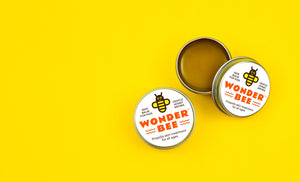 Two Wonder Bee Skin Treatment tins on a yellow background. One is open to reveal the contents.